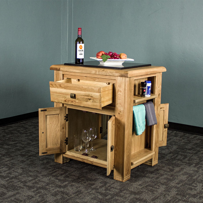 The front of the Danube Compact Granite Top Oak Kitchen Island with its doors and drawers open, there is a fruit platter and a bottle of wine on the granite top, and two towels hanging on the side towel rack, with salt and pepper on the small shelf above it. There are two sets of glasses and wine glasses in the middle shelf/opening.