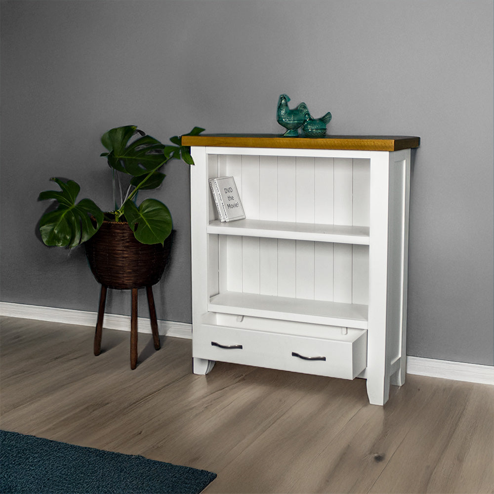 The front of the Felixstowe 1m Bookcase with its drawers open. There are two DVDs on the top shelf and two blue glass ornaments on top of the bookshelf. There is a free standing potted plant next to it.