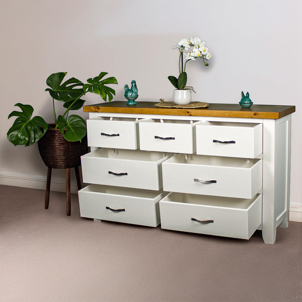 The front of the Felixstowe 7 Drawer Pine Lowboy with its drawers open. There are two blue glass ornaments on top with a small pot of white flowers on a rattan leaf in between them. There is a free standing potted plant next to the lowboy.