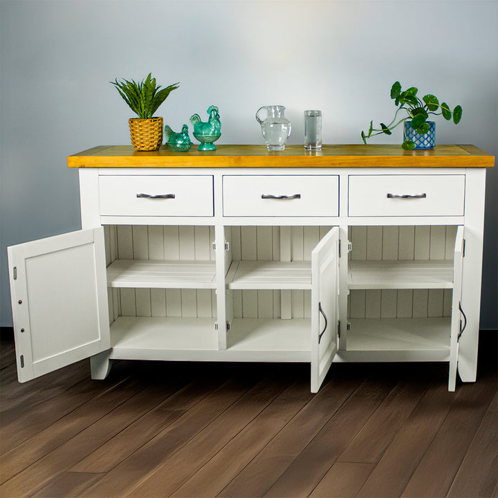 The front of the Felixstowe Large Pine White Buffet with its doors open. From left to right: a potted plant, two blue glass ornaments, a water pitcher, a glass of water and a potted plant are all on top of the sideboard.
