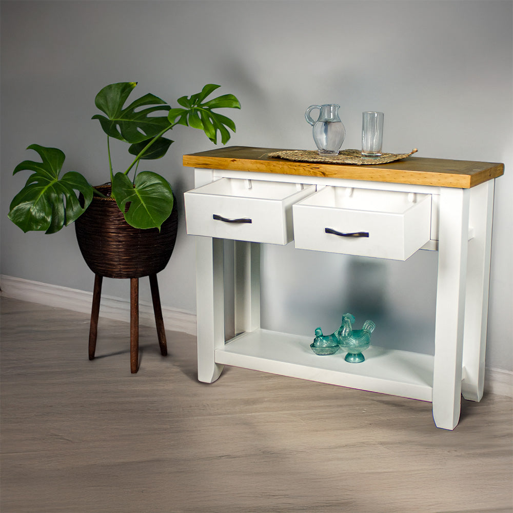 The front of the Felixstowe Large Pine Hall Table with its drawers open. There are two blue glass ornaments on the lower shelf, and a glass water pitcher and glass of water on a rattan leaf on top of the console table. There is a free standing potted plant next to the hallway table.