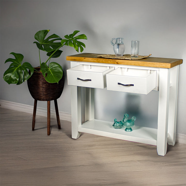 The front of the Felixstowe Large Pine Hall Table with its drawers open. There are two blue glass ornaments on the lower shelf, and a glass water pitcher and glass of water on a rattan leaf on top of the console table. There is a free standing potted plant next to the hallway table.