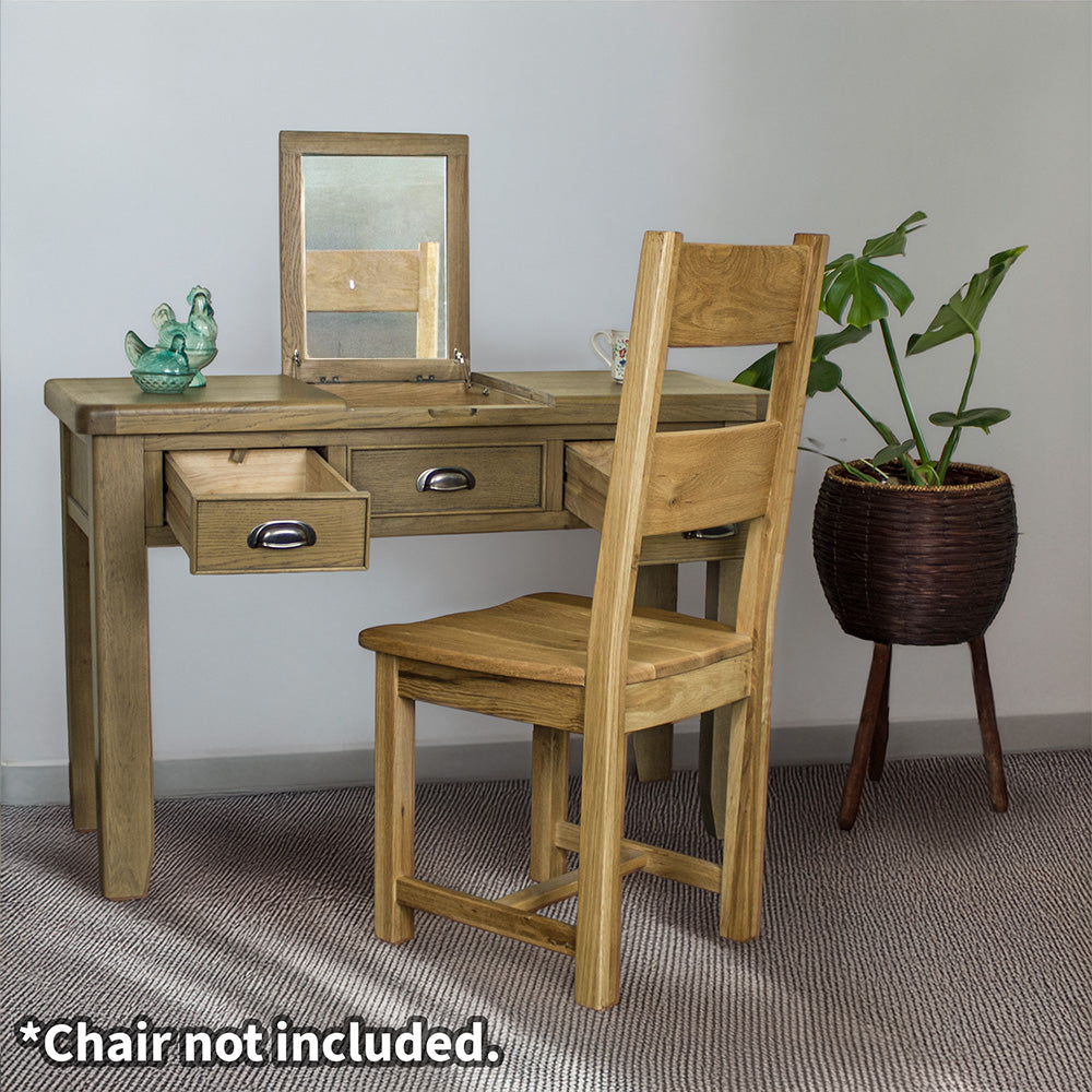 The front of the Houston Oak Dressing Table With Mirror with its drawers open. There is an oak dining chair in front. There are two blue glass ornaments on top of the table and a free standing potted plant to the right of the desk.
