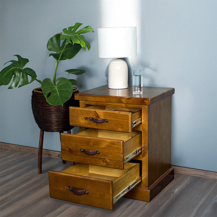 The front of the Jamaica Bedside Cabinet with its drawers open. There is a lamp and glass of water on top. There is a potted plant next to the bedside table.