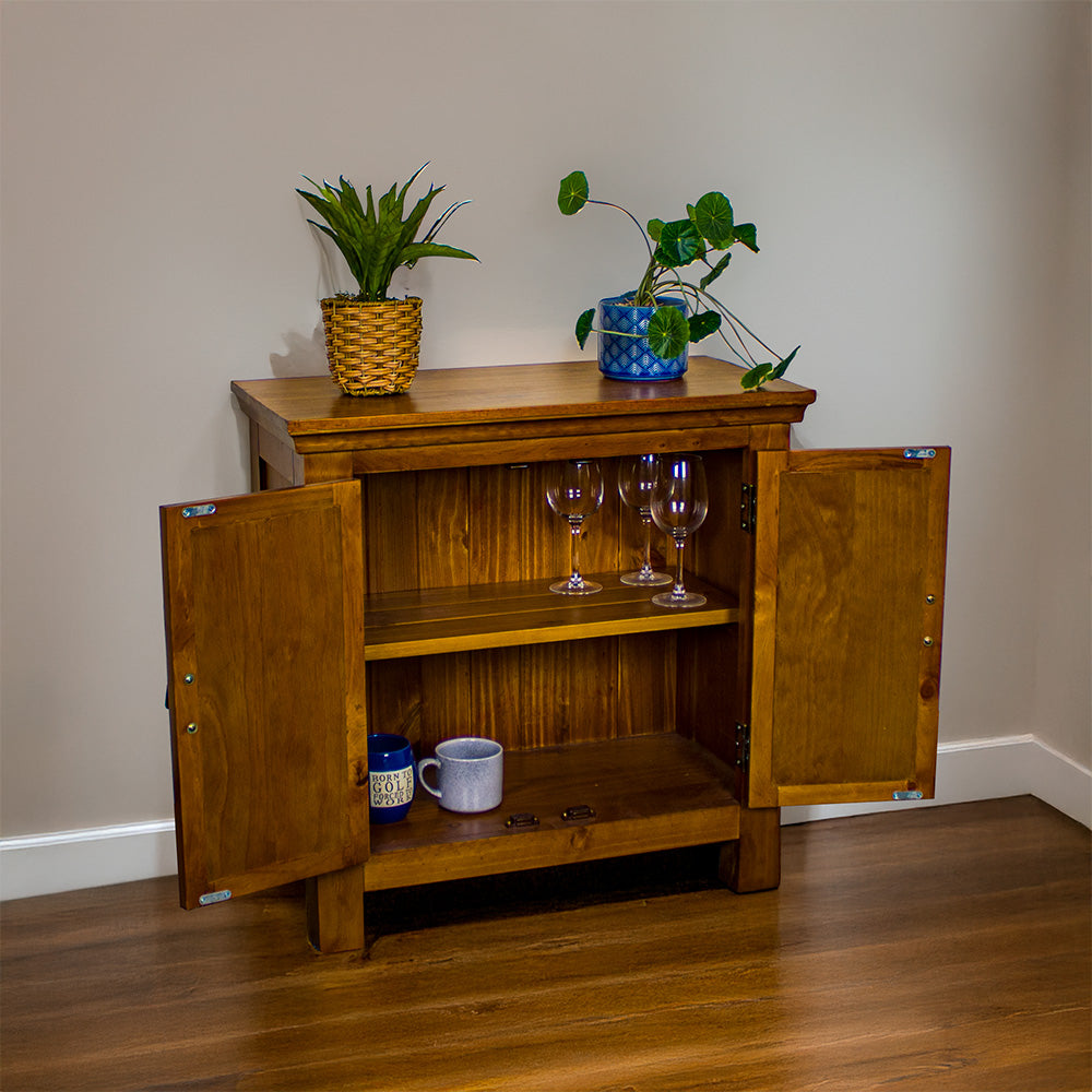 The front of the Montreal Compact Pine Buffet with its doors open. There are two potted plants on top. There are three wine glasses on the top shelf and two coffee mugs on the lower shelf.