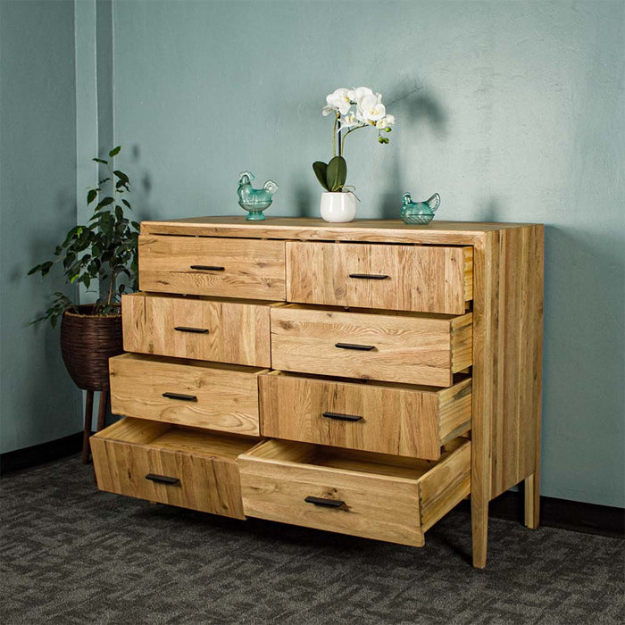 The front of the Ormond 8 Drawer Oak Tallboy with its drawers open. There is a free standing potted plant next to it. There is a small pot of white flowers on top in the middle with two blue glass ornaments on either side.