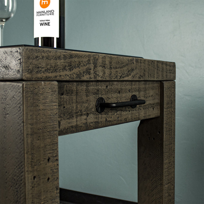 Side of the Stonemill Recycled Pine Granite Workbench, there is a black metal handle. The side of a glass and the lower half of a glass bottle can be seen.