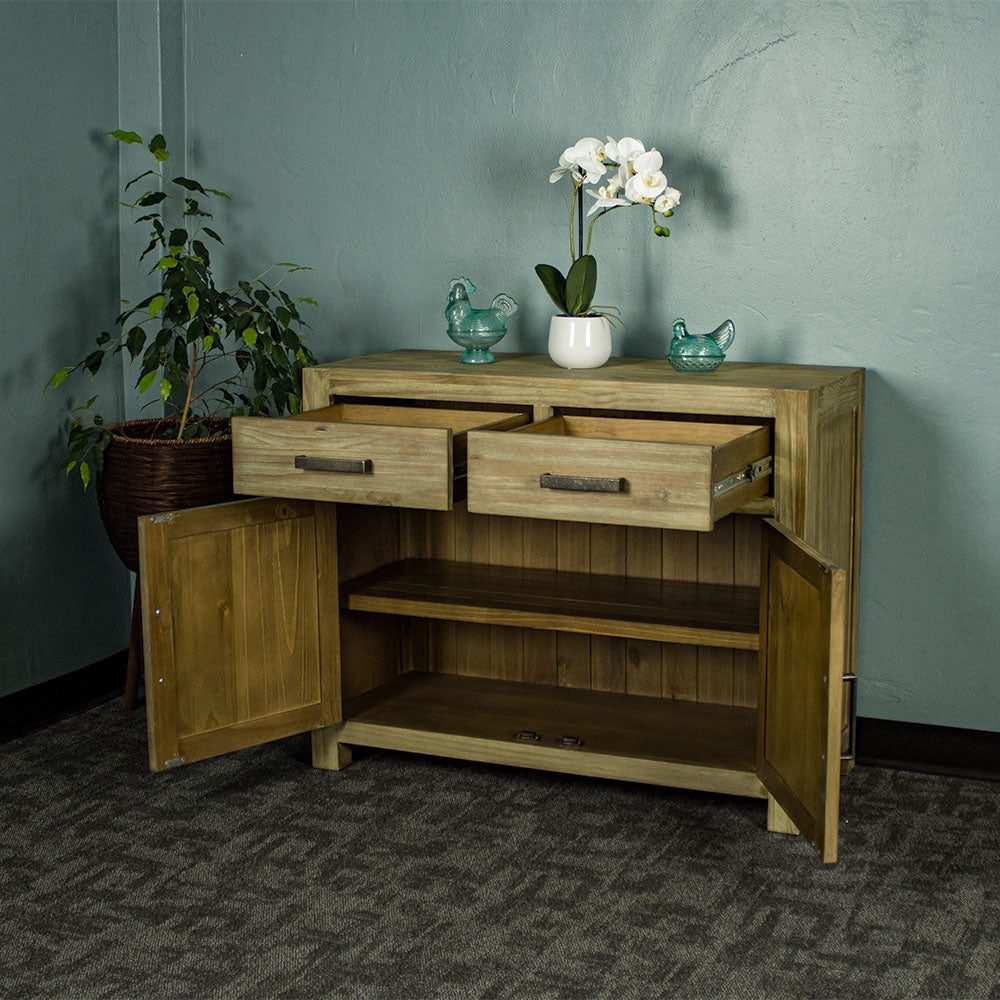 The front of the Vancouver Pine Buffet with its doors and drawers open. There are two blue glass ornaments on top with a pot of white flowers in between. There is a free standing potted plant next to the buffet.