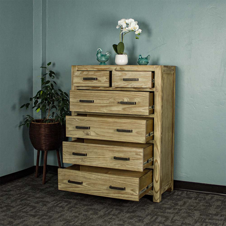 The front of the Vancouver 6 Drawer NZ Pine Tallboy with its drawers open. There are two blue glass ornaments on top with a small pot of white flowers in between. There is a free standing potted plant next to the unit.