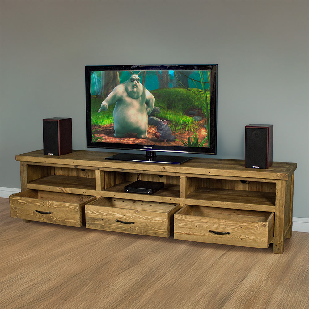 The front of the Ventura Recycled Pine Large TV Unit with its drawers open. There are two speakers on either side of a large TV in the middle on top of the unit. There is a DVD player in the middle shelf. There is a free standing tall potted plant next to the unit.