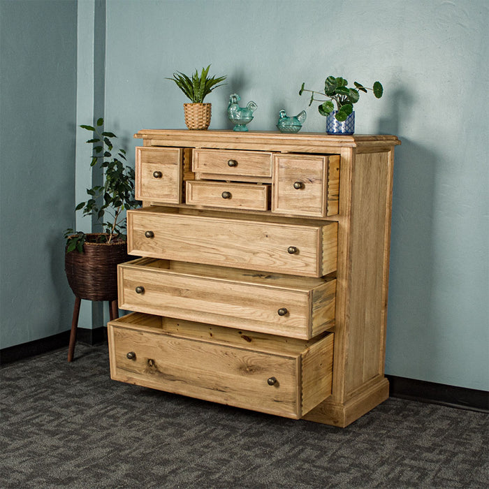 The front of the Versailles Oak 7 Drawer Chest with its drawers open. There are two potted plants and two blue glass ornaments on top. There is a free standing potted plant next to it.