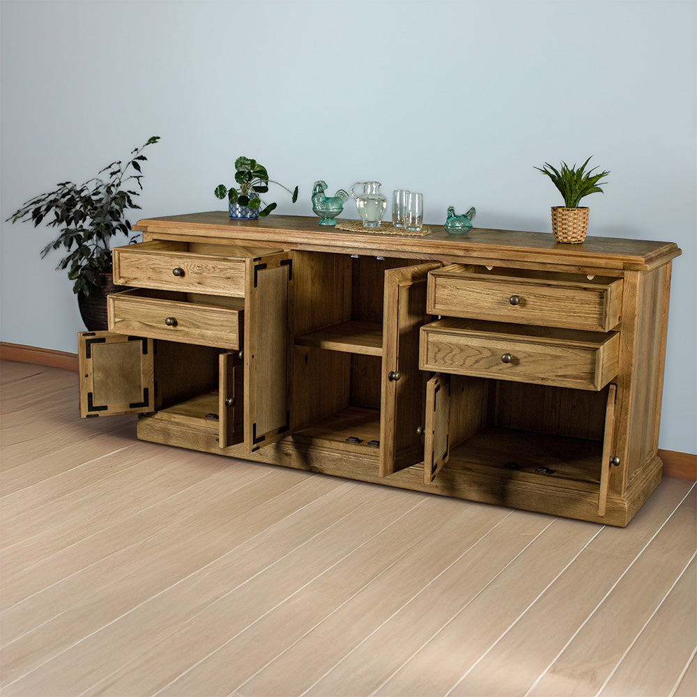 The front of the Versailles Large Oak Buffet with its drawers and doors open. There are two potted plants on either side, and two blue glass ornaments, a pitcher of water next to two glasses on a rattan style leaf coaster in the middle on top.