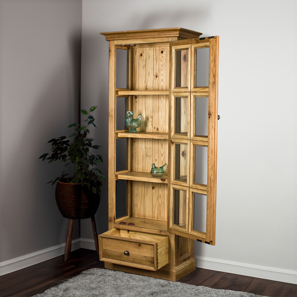The Versailles Small Display Cabinet with its door and drawer open. There are two blue glass ornaments on the middle two shelves. There is a tall, free standing potted plant next to the cabinet.