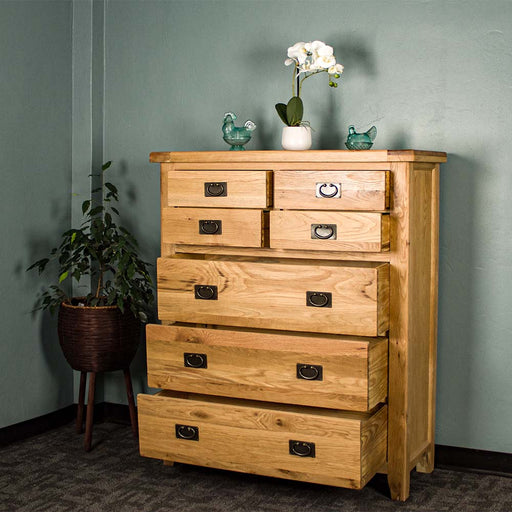The front of the Vienna 7 Drawer Oak Tallboy with its drawers open. There are two blue glass ornaments on top with a pot of white flowers in between. There is a free standing potted plant next to the unit.