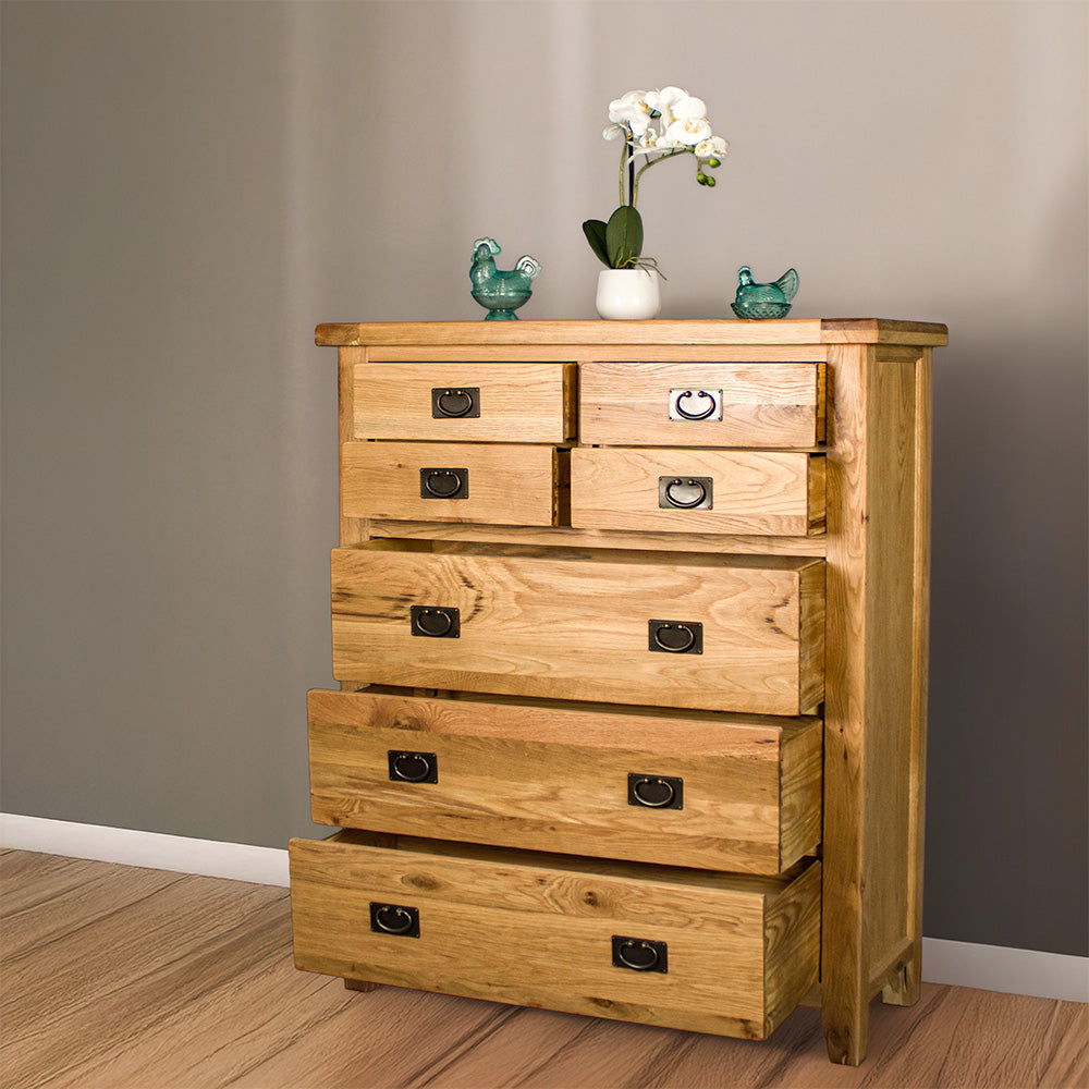 The front of the Vienna 7 Drawer Oak Tallboy with its drawers open. There are two blue glass ornaments on top with a pot of white flowers in between. 