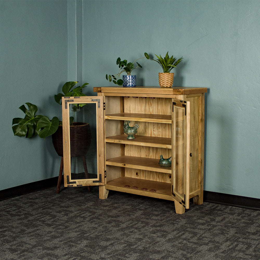 The front of the Yes Oak Display Cabinet with its doors open. There are two blue glass ornaments on the middle shelves. There are two potted plants on top and a tall, free standing potted plant next to the cabinet.