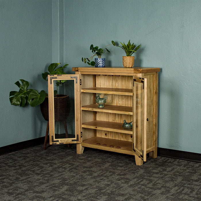 The front of the Yes Oak Display Cabinet with its doors open. There are two blue glass ornaments on the middle shelves. There are two potted plants on top and a tall, free standing potted plant next to the cabinet.