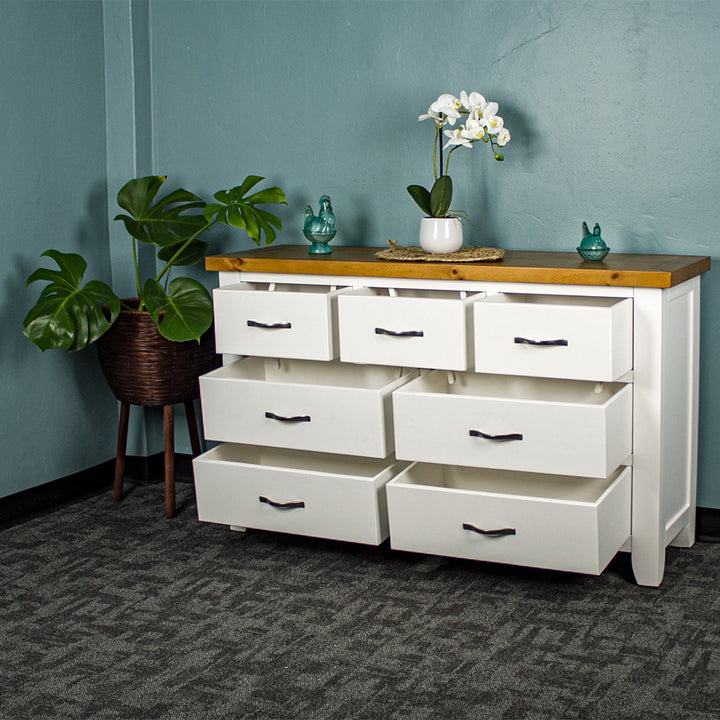 The front of the Felixstowe 7 Drawer Pine Lowboy with its drawers open. There are two blue glass ornaments on top with a small pot of white flowers on a rattan leaf in between them. There is a free standing potted plant next to the lowboy.