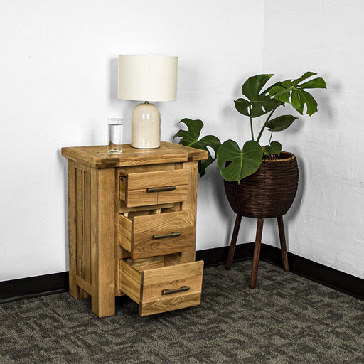 An overall view of the Camden 3 Drawer White Oak Bedside Table with its drawers open. There is a glass of water and a lamp on top. There is a free standing potted plant next to the bedside table.