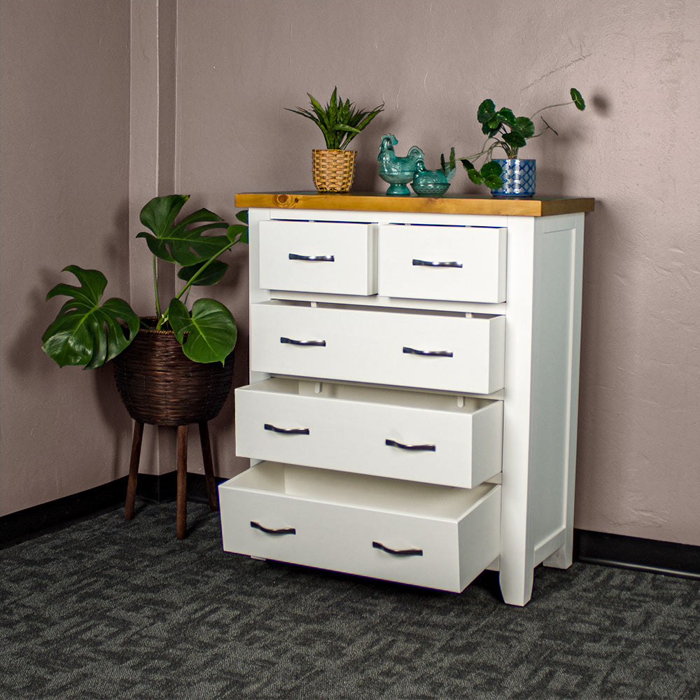 The front of the Felixstowe 5 Drawer Pine Tallboy, with its drawers open. There are two potted plants on top with two blue glass ornaments in between. There is a free standing potted plant next to the tallboy.