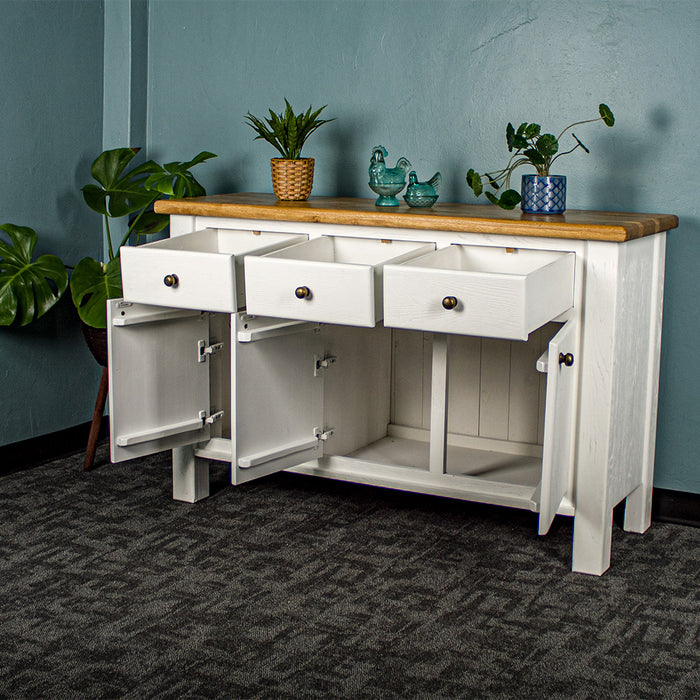 The front of the Loire Two-Tone Medium Oak Buffet with its drawers and doors open. There are two potted plants on top with two blue glass ornaments in between. There is a free standing potted plant to the left of the sideboard.