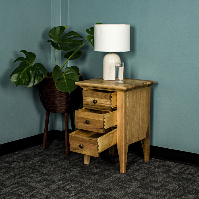 The Beethoven 3 Drawer Oak Bedside Cabinet with its drawers open. There is a glass of water and a white lamp on top and a large free standing potted plant next to it.