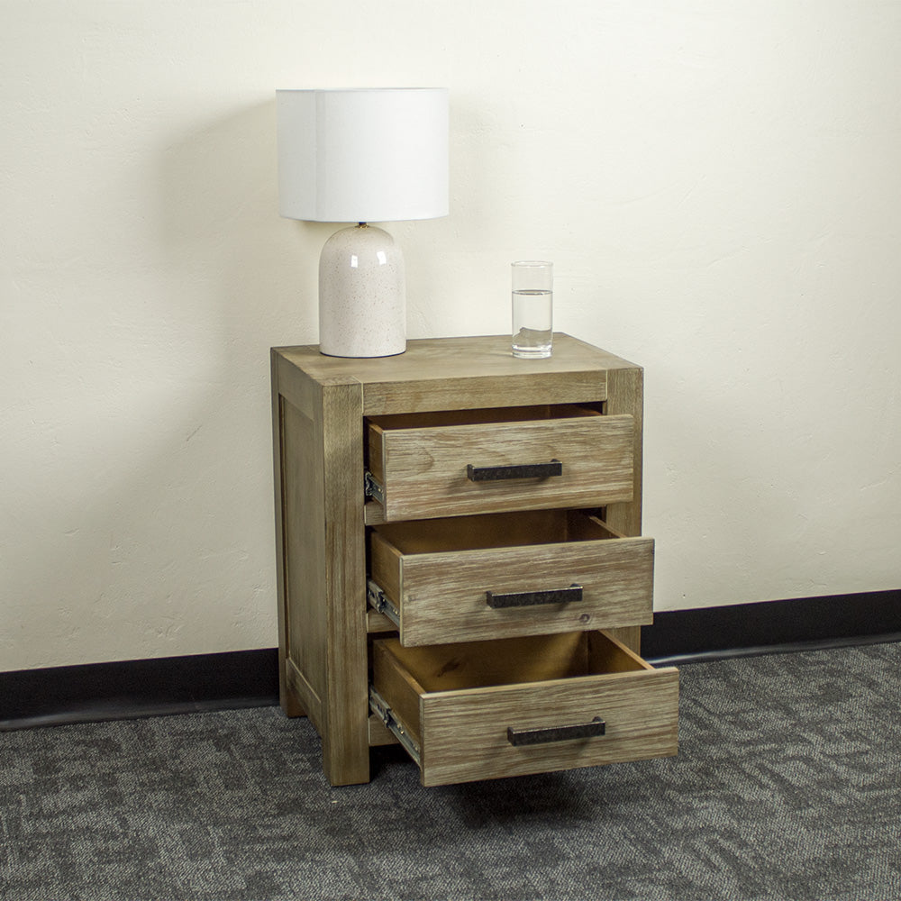 The front of the stonewash Vancouver 3 Drawer Bedside Cabinet with its drawers open. There is a lamp and a glass of water on top.