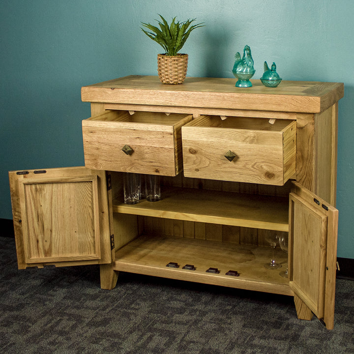 The front of the Maximus Small Oak Buffet with its drawers and doors open. There is a potted plant and two blue glass ornaments on top. There are three glasses on the top shelf, and two wine glasses on the bottom shelf.