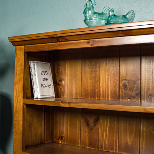 A closer view of the top shelf of the Montreal Short Pine Bookcase. There are two DVD cases on the shelf and two blue glass ornaments in the shape of chickens on top of the bookshelf.