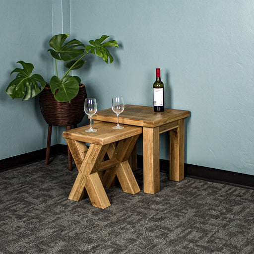 An overall view of the Amstel Oak Nesting Tables. The smaller table is out from underneath the top one. There is a free standing potted plant next to the tables. There are two wine glasses on top of the smaller table and a bottle of wine on the larger table.