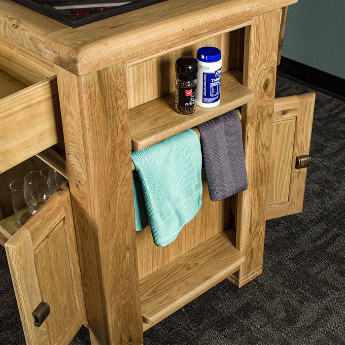 The side of the Danube Compact Granite Top Oak Kitchen Island, showing off the towel rack, which has a blue towel and a purple towel hanging on it. There is salt and pepper shakers on the small shelf above the towel rack.