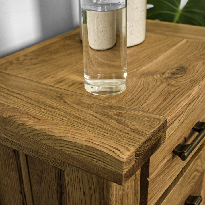 A close up of the top of the Camden 3 Drawer White Oak Bedside Table, showing the wood grain. There is a glass of water on top.