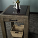 Full side of the Stonemill Recycled Pine Granite Workbench, showing the symmetry of both sides. The black metal handle is on display in the front, and the black metal hooks can be seen. There is a woven basket on the lower shelf. There are two glasses on the granite top and a glass bottle in front of them.