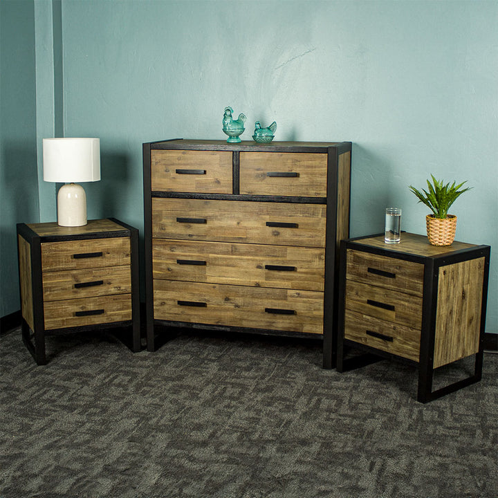 The Victor 7 Drawer Tallboy with two Victor 3 Drawer Bedsides on either side. There are two blue glass ornaments on top of the tallboy, a lamp on the right bedside and a glass of water and potted plant on the right bedside.