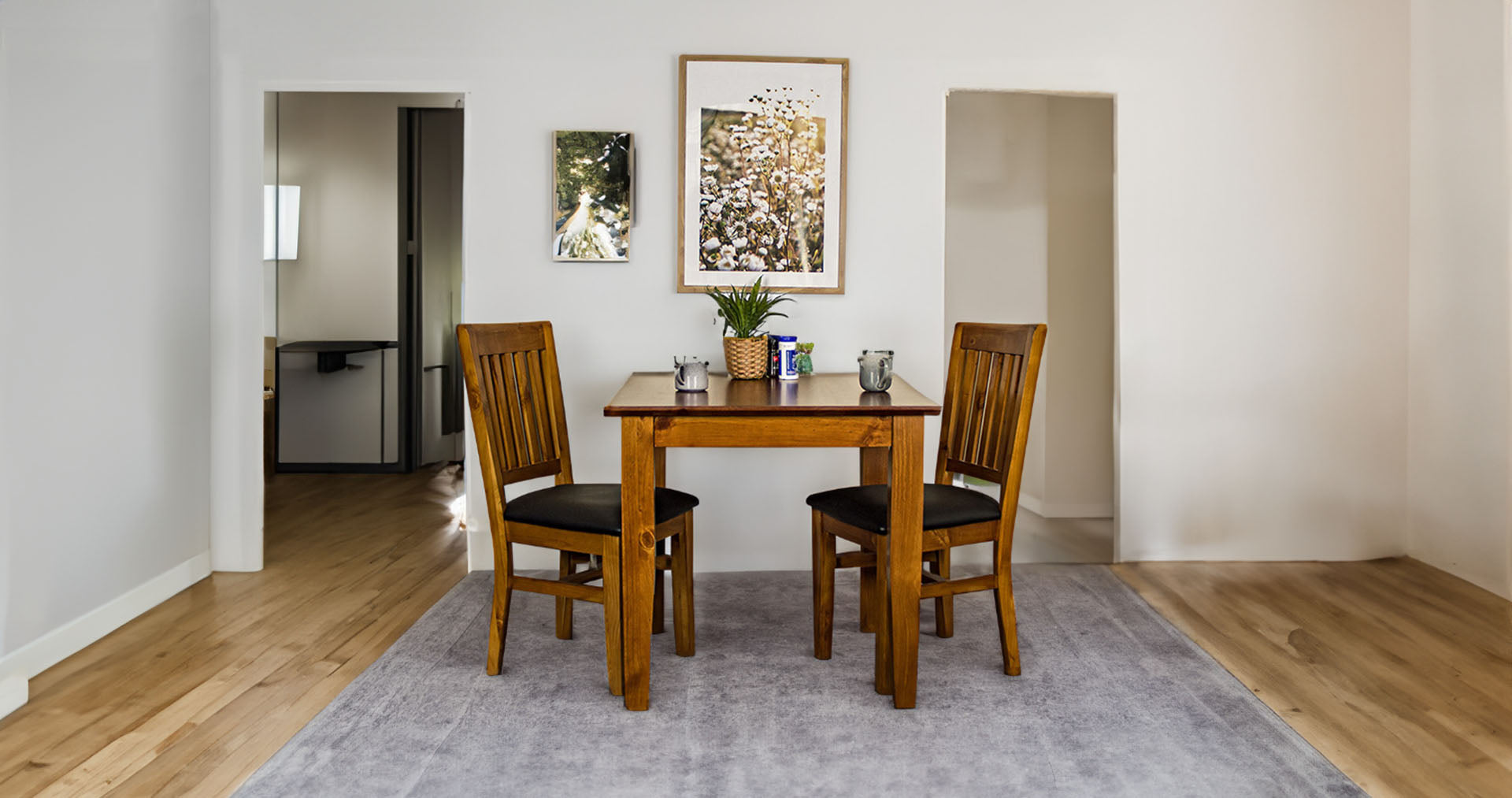 Dining room with light gray walls and wooden floorboards. There is a large gray rug in the middle of the room with a Hamilton 3 PIece Dining Suite on top, with a Hamilton Square Rimu Stained Dining Table, which has two coffee mugs, a potted plant and a salt and pepper shaker on top. There are two Hamilton Rimu-stained upholstered Dining Chairs on opposite ends of the dining table. There are two framed artworks on the back wall above the dining table and chairs.