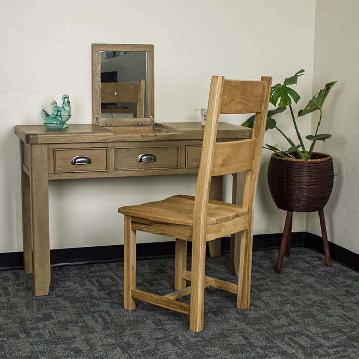 The front of the Houston Oak Dressing Table With Mirror. There is an oak dining chair in front. There are two blue glass ornaments on top of the dressing table and a free standing potted plant to the right of the table.