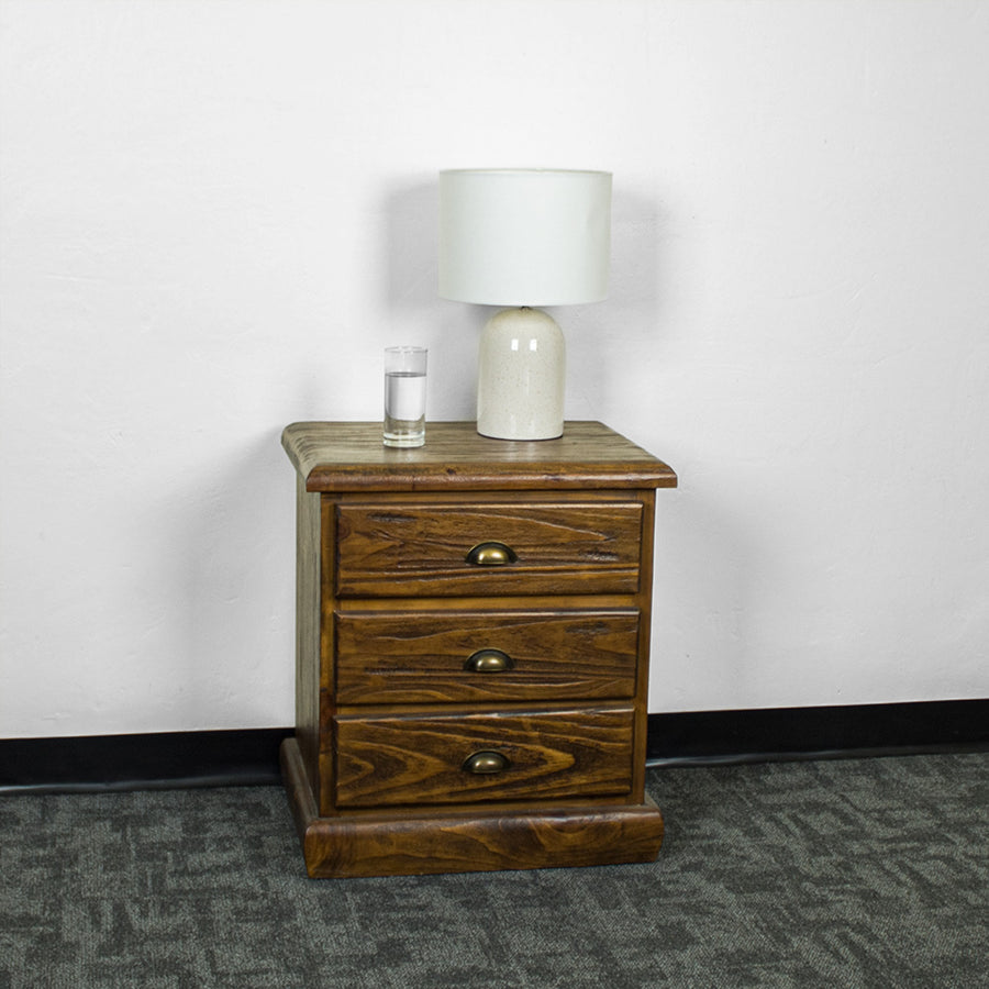 The front of the Heid Large 3 Drawer Pine Bedside. There is a lamp and a glass of water on top.