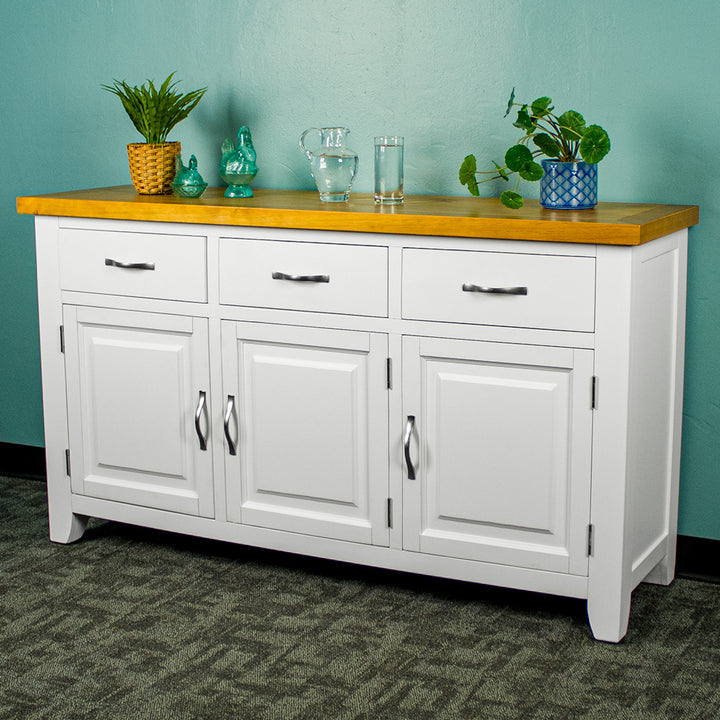 The front of the Felixstowe Large Pine White Buffet. From left to right there are: a potted plant, two blue glass ornaments, a water pitcher, a glass of water and a potted plant on top of the sideboard.