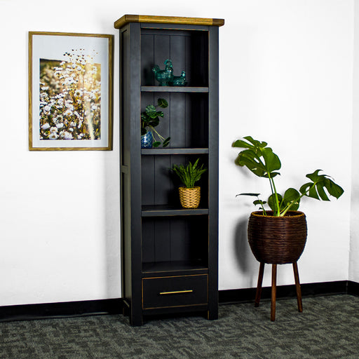 The front of the Cascais Tall Black Bookcase. There is a framed photo of flowers on the wall on the left and a tall, free standing potted plant on the right on the ground. There are two blue glass ornaments on the top shelf, a potted plant on the second shelf, and a third on the bottom shelf.
