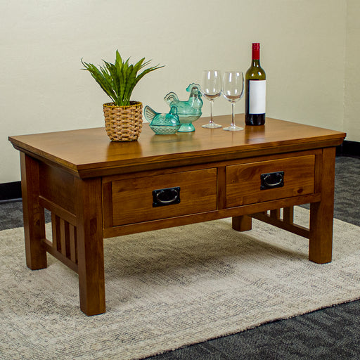 The front of the Montreal Coffee Table with 2 Drawers. There is a potted plant, two blue glass ornaments, two wine glasses, and a bottle of wine on top.