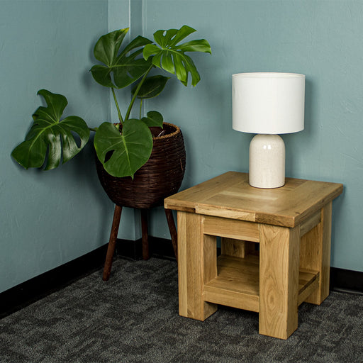 An overall view of the Camden Oak Lamp Table. There is a lamp on top and a free standing potted plant to the side.