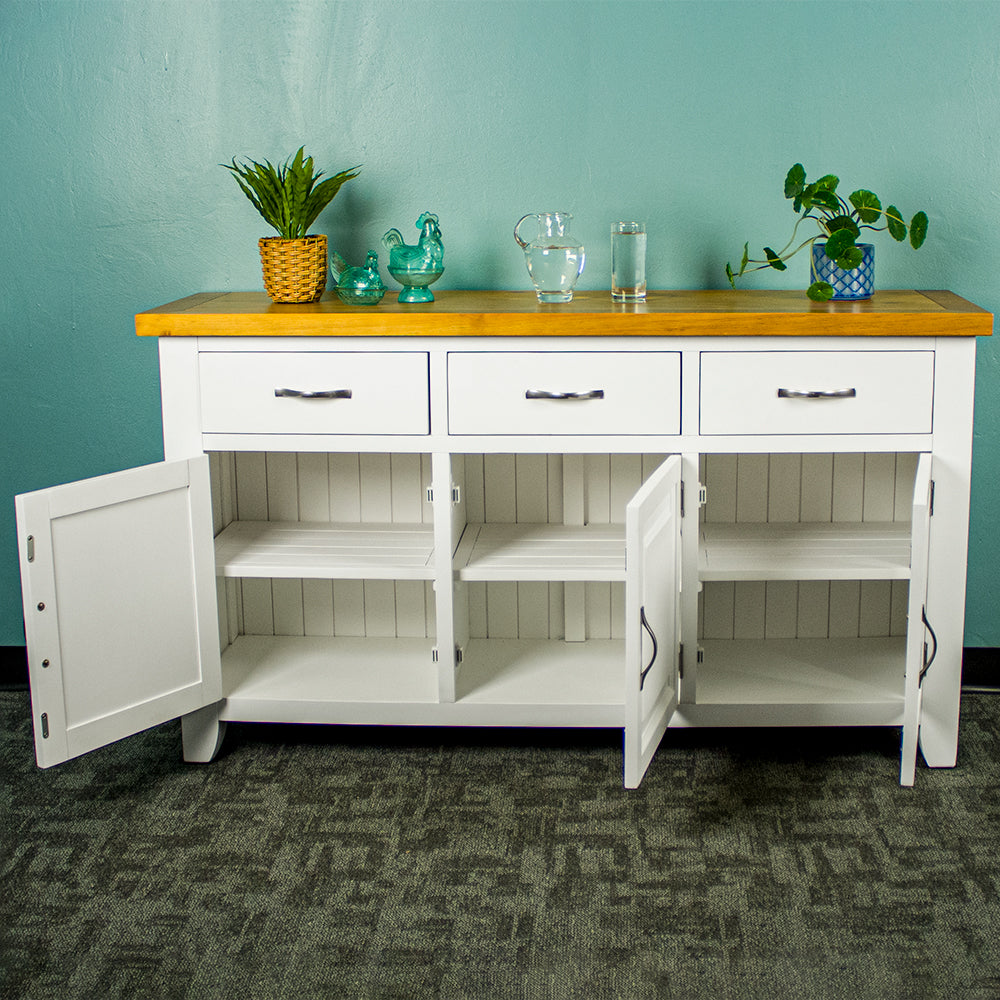 The front of the Felixstowe Large Pine White Buffet with its doors open. From left to right: a potted plant, two blue glass ornaments, a water pitcher, a glass of water and a potted plant are all on top of the sideboard.