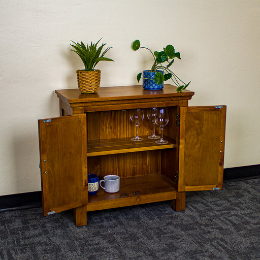 The front of the Montreal Compact Pine Buffet with its doors open. There are two potted plants on top. There are three wine glasses on the top shelf and two coffee mugs on the lower shelf.
