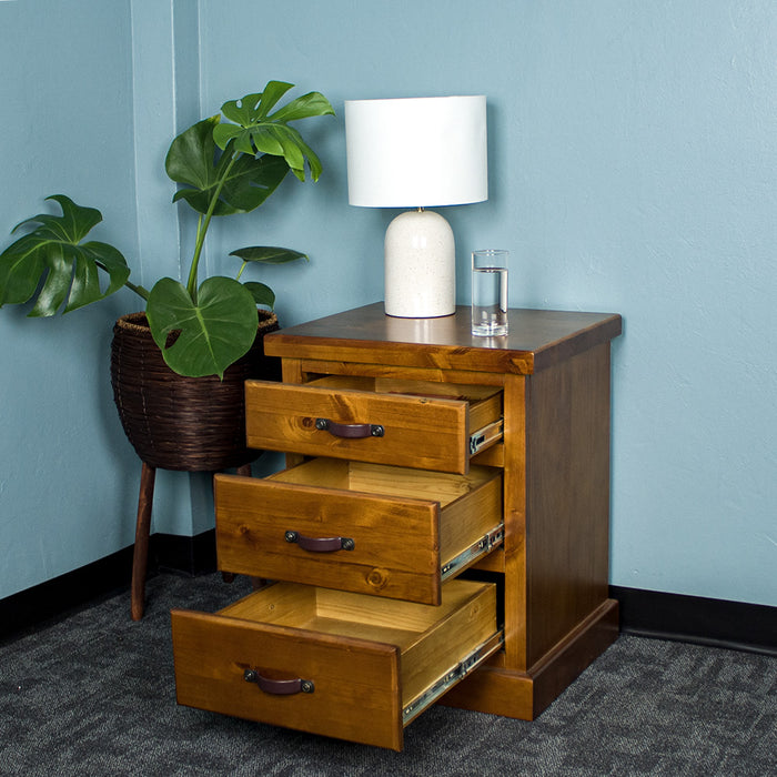 The front of the Jamaica Bedside Cabinet with its drawers open. There is a lamp and a glass of water on top. There is a potted plant next to the bedside table.