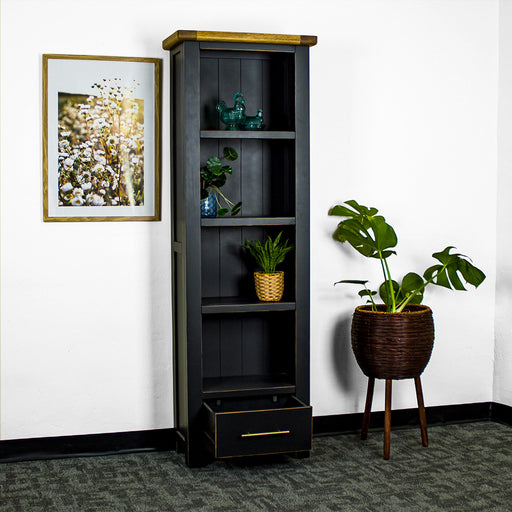 The front of the Cascais Tall Black Bookcase with its drawer open. There is a framed photo of flowers on the wall on the left and a tall, free standing potted plant on the right on the ground. There are two blue glass ornaments on the top shelf, a potted plant on the second shelf, and a third on the bottom shelf.