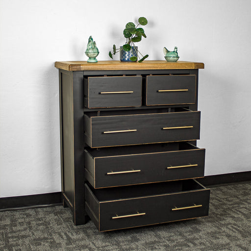 The front of the Cascais Oak Top Black Tallboy with its drawers open. There is a potted plant on top in the middle with two blue glass ornaments on either side.