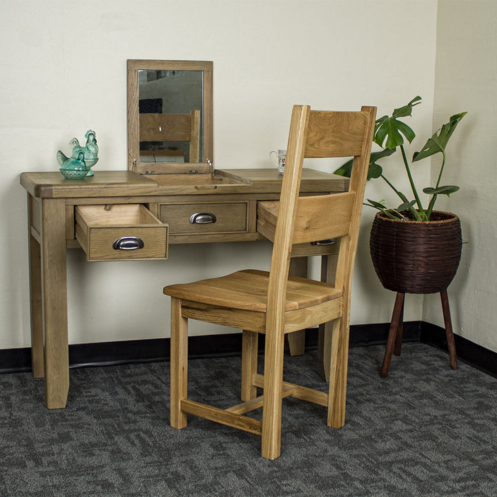 The front of the Houston Oak Dressing Table With Mirror with its drawers open. There is an oak dining chair in front. There are two blue glass ornaments on top of the table and a free standing potted plant to the right of the desk.