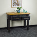 The front of the Cascais 2-Drawer Hall Table with its drawers open. There is a potted plant on top of a rattan leaf in the middle, with two blue glass ornaments next to it on top. There is a framed picture of flowers on the wall.