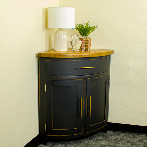 The front of the Cascais Oak-Top Corner Cabinet.  There is a lamp, a water pitcher, a glass and a potted plant on top.