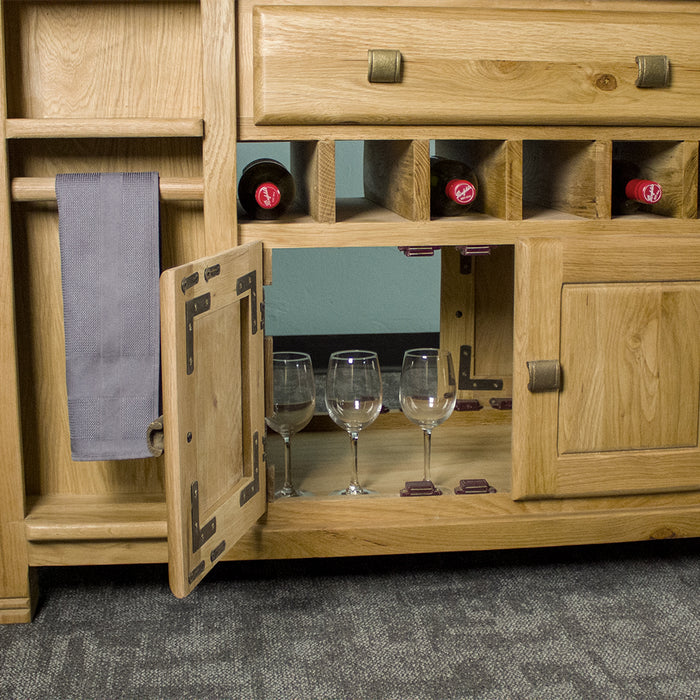 A closer view of the Danube Large Granite Top Oak Kitchen Island's open ended lower shelf. There are three wine glasses inside. There are three wine bottles on the small wine rack above and a hand towel hanging on the towel rail to the left.